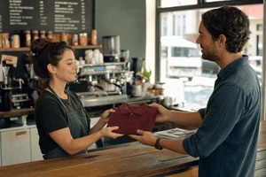 A cafe owner hands a folded blank burgundy polo shirt to a staff member behind a counter