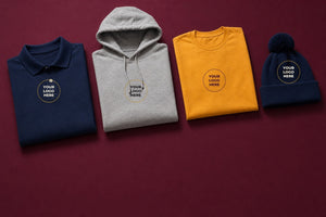 Folded blank custom apparel, a navy polo, grey pullover hoodie, gold t-shirt, and navy pom beanie, each marked with a Your Logo Here placeholder emblem, on a mulberry background.