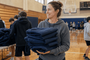 A youth sports coach holds a stack of folded blank navy hoodies in a school gymnasium