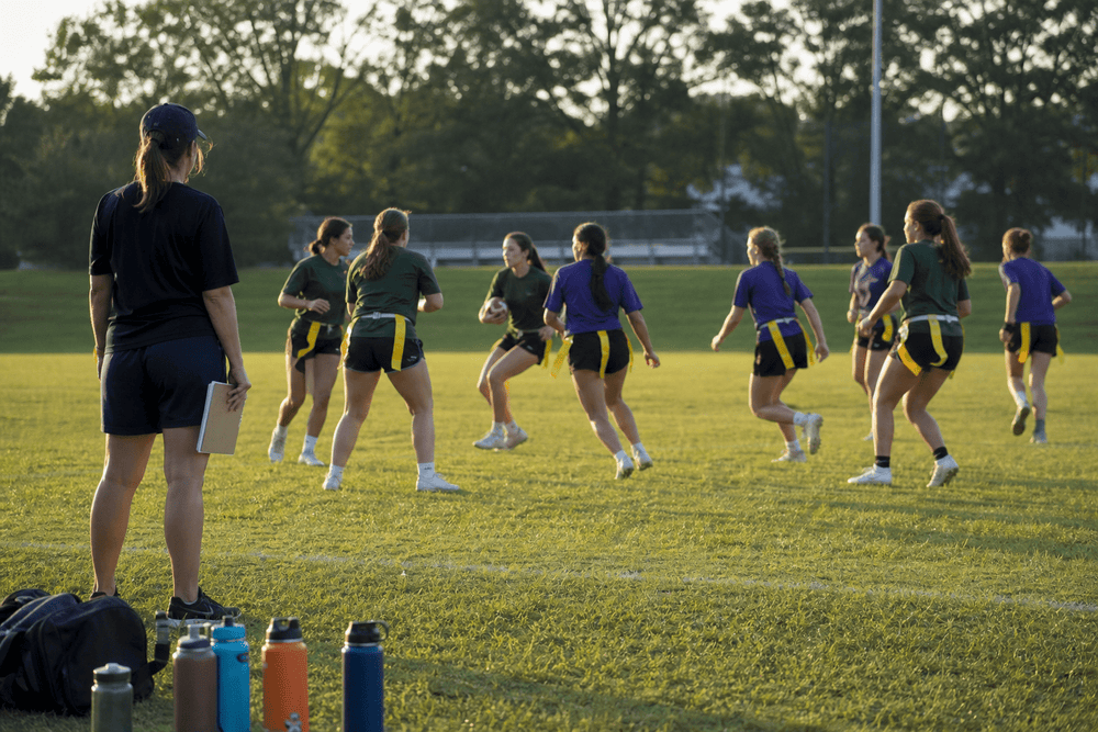 High school girls flag football team practicing on a grass field at sunset while a coach watches from the sideline