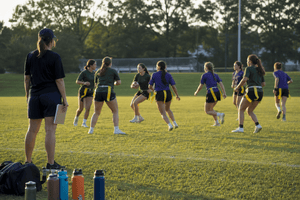 High school girls flag football team practicing on a grass field at sunset while a coach watches from the sideline