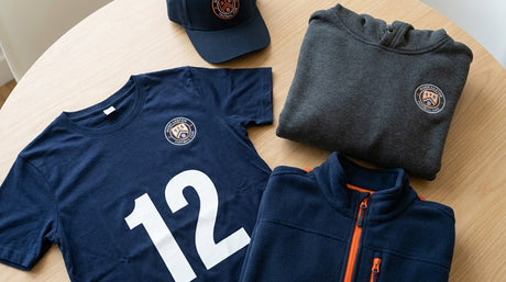 Flat lay of custom navy youth team apparel: a number 12 jersey, embroidered shield cap, crest hoodie, and orange-zip fleece