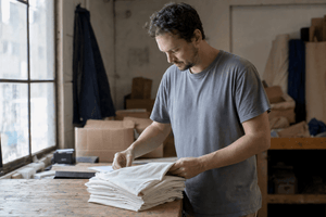 Man inspecting a stack of blank undyed cotton t-shirts on a wooden workroom table