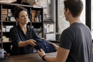 Small business owner handing a folded navy polo shirt to a new employee behind a shop counter