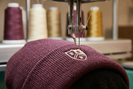 Embroidery machine needle stitching a tonal logo onto the cuff of a mulberry knit beanie